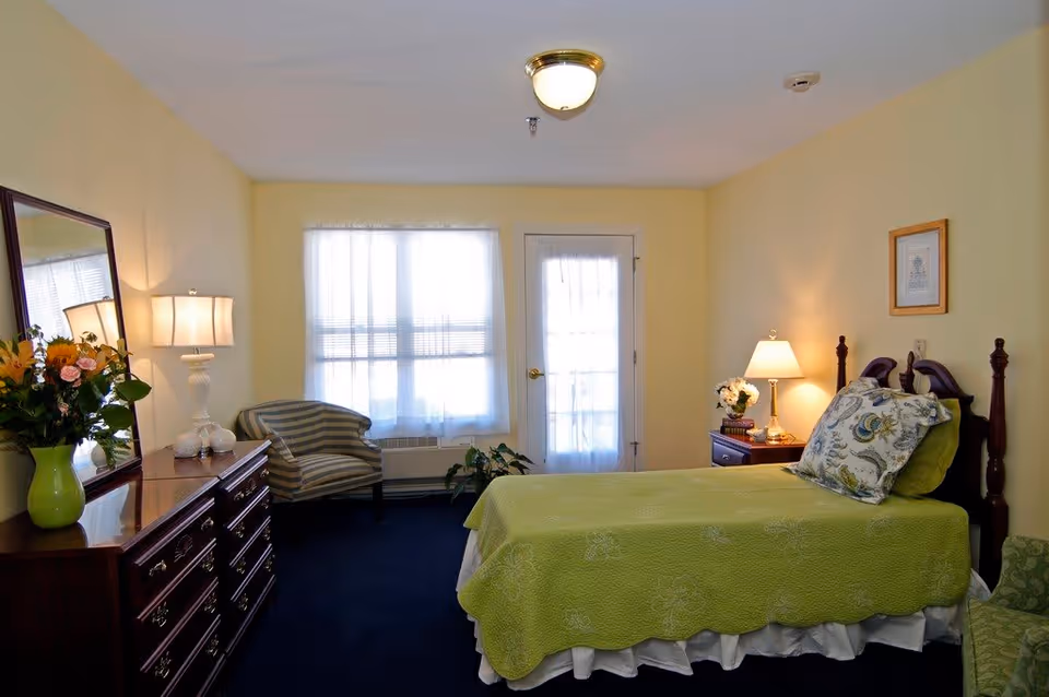 A cozy bedroom with a single bed covered in a green quilt and floral pillows. The room has light yellow walls, a wooden dresser with a mirror and a vase of flowers, a striped armchair near a window with sheer curtains, a nightstand with a lamp and decorative items, and a door with a glass panel.