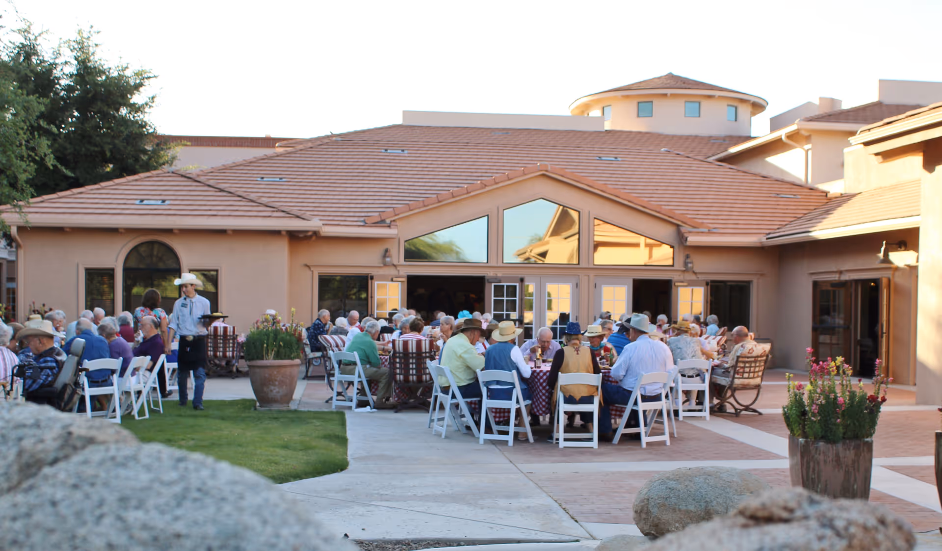 Outdoor patio area at La Posada at Green Valley with multiple tables where elderly people are seated and socializing. A staff member wearing a cowboy hat is walking among the tables. The building has a tan exterior with a tiled roof and large windows.