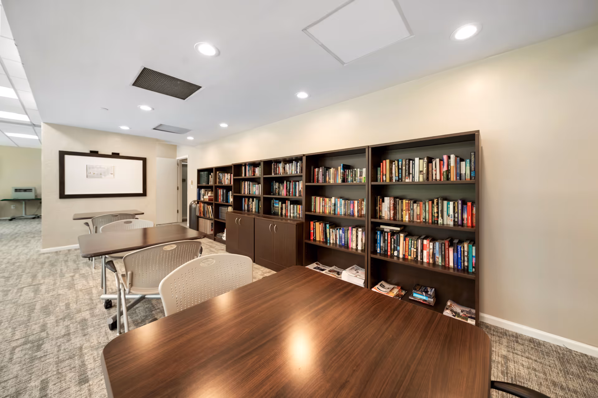 Interior room with several wooden tables and beige chairs arranged in rows. Along the right wall, there are multiple dark wooden bookshelves filled with books and some magazines on the bottom shelves. The room has a light-colored carpet and ceiling lights.