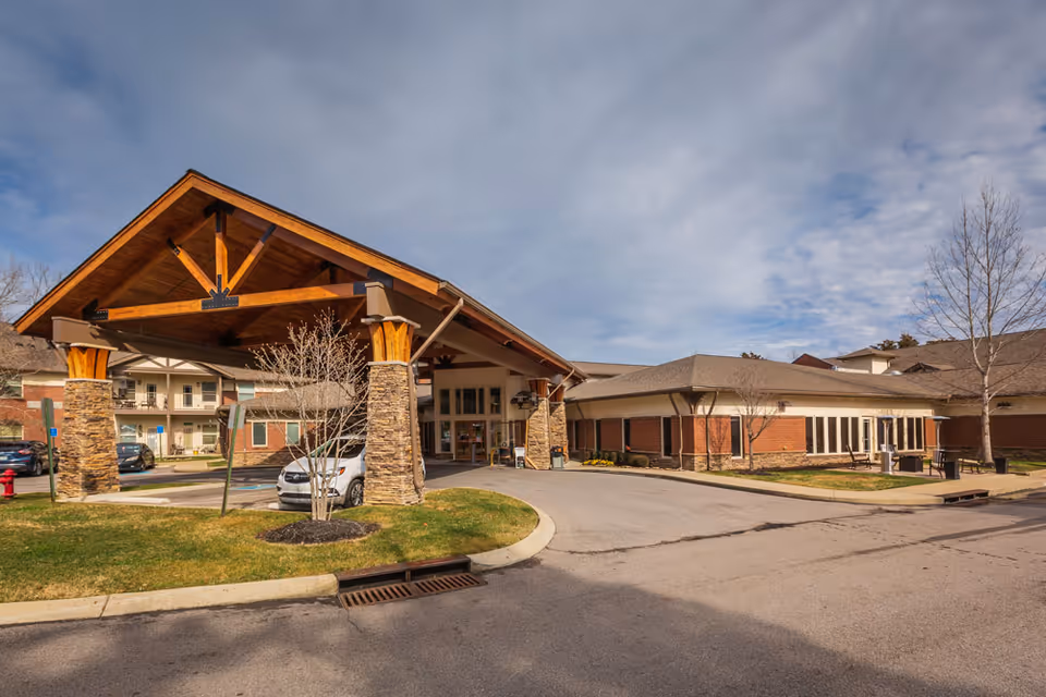 Exterior view of The Lodge at Natchez Trace senior living facility showing a large covered entrance with wooden beams and stone pillars, a driveway, parked cars, and a partly cloudy sky.