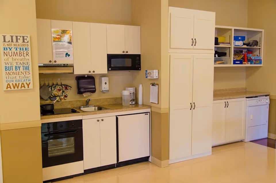 A clean and organized kitchen area with beige walls and white cabinets. The kitchen includes an oven, microwave, sink, dishwasher, and refrigerator. There are kitchen utensils hanging on the wall, a water pitcher, paper towels, and a coffee maker on the countertop. On the left wall, there is a decorative sign with an inspirational quote about life.
