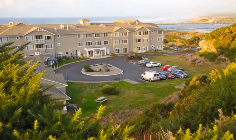 Three-story senior living building by the coast with a circular driveway, parked cars, and surrounding grassy landscaping.