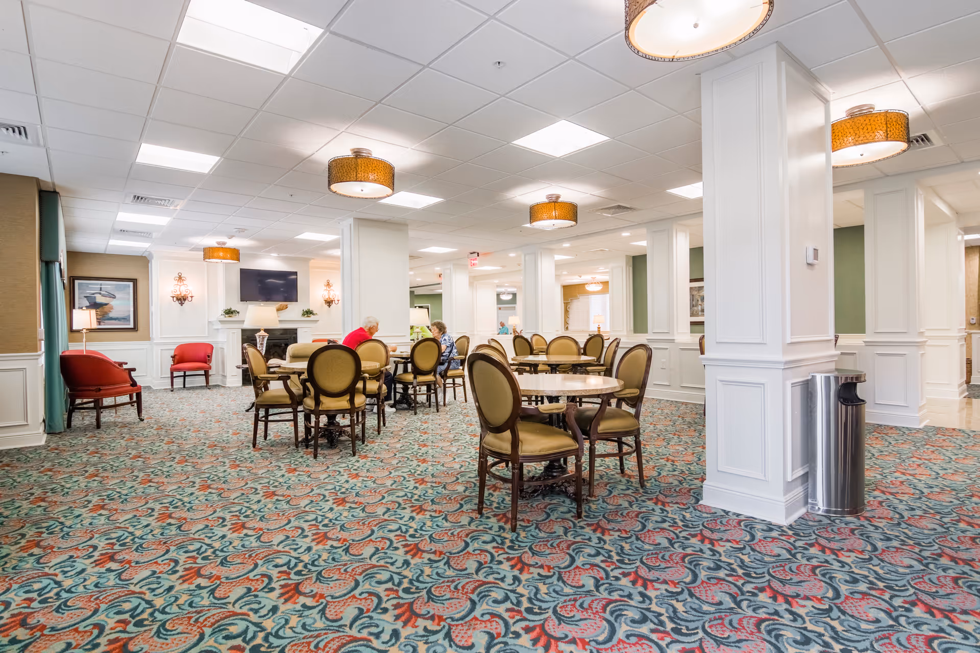 A spacious, well-lit common area in a senior living facility with patterned carpet, round tables with chairs, and a few seniors seated and conversing. The room features white paneled columns, wall sconces, a fireplace with a TV above it, and decorative ceiling lights.
