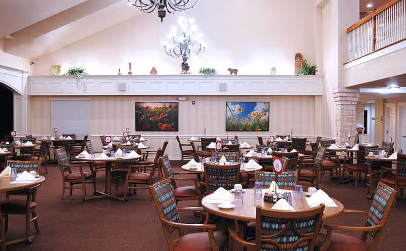 Large dining room with round tables set for service, patterned chairs, chandeliers, and decorative wall art.