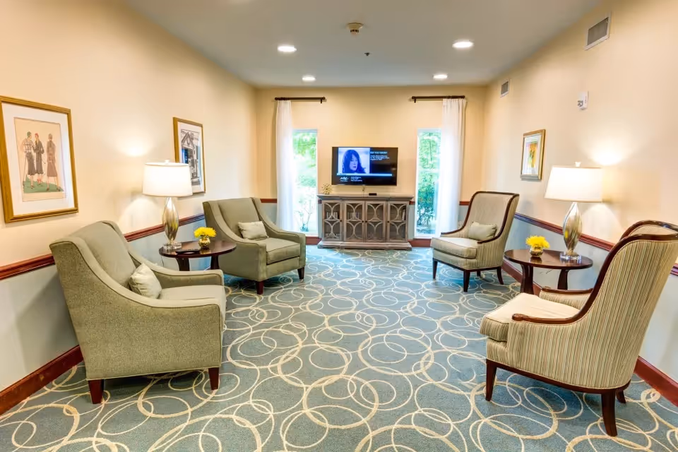 A cozy senior living facility lounge with four armchairs arranged around two small round tables, each table holding a lamp and a small vase with yellow flowers. The room has light beige walls adorned with framed artwork, a patterned carpet with circular designs, and a flat-screen TV mounted on a cabinet between two tall windows with white curtains.