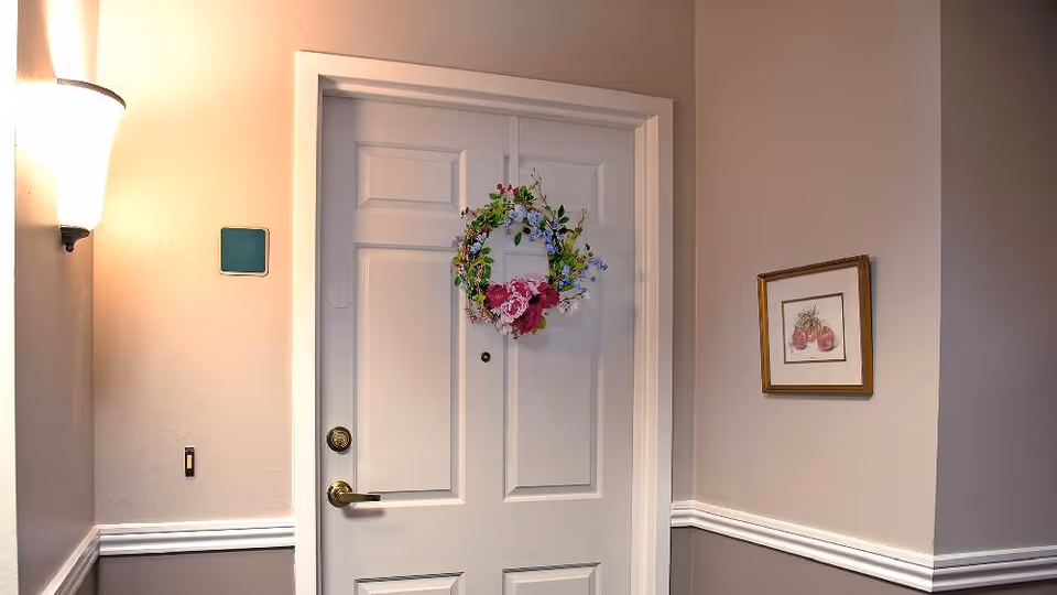 A white door decorated with a colorful floral wreath hanging in the center. The door has a brass handle and a peephole. The surrounding walls are painted beige with white trim, and there is a wall-mounted light fixture on the left side and a framed picture of tomatoes on the right wall.