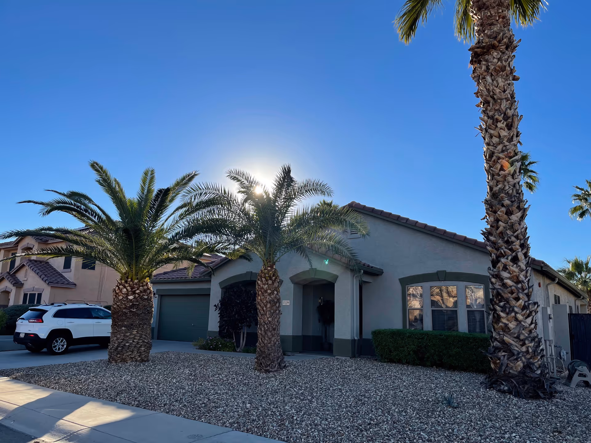 Exterior view of a single-story residential building with a tiled roof, three palm trees in the front yard covered with gravel, a white SUV parked in the driveway, and clear blue sky with the sun shining behind the central palm tree.