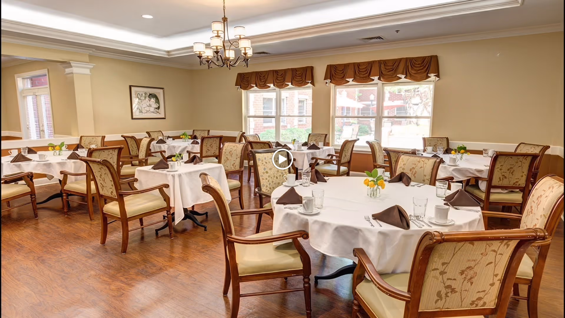 Bright dining room with round tables set with white tablecloths, wooden chairs, place settings, and a chandelier.