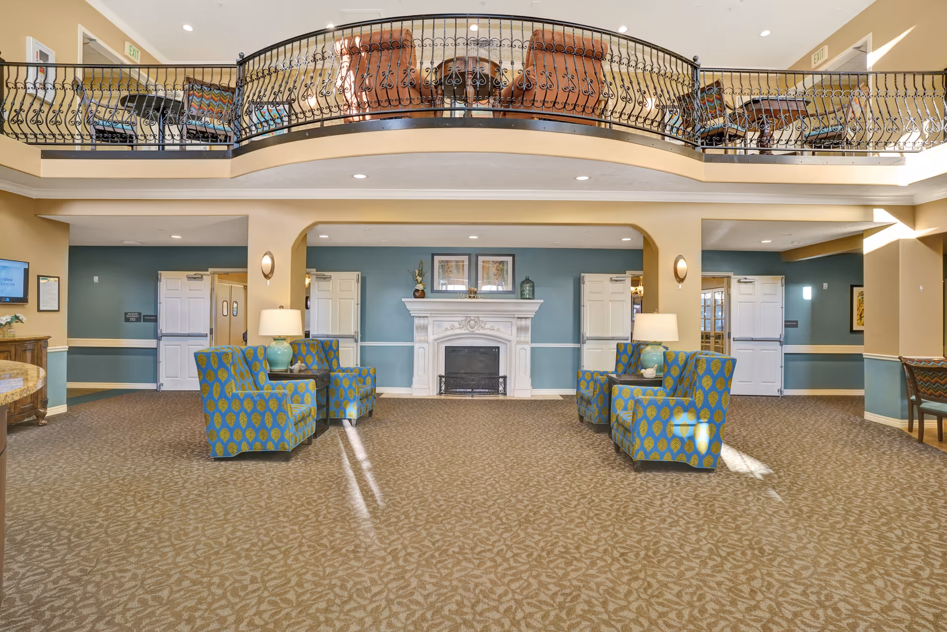 A spacious senior living facility common area with patterned carpet and beige walls. The room features a white decorative fireplace centered on a blue accent wall, flanked by two pairs of blue and green patterned armchairs with side tables and lamps. Above, there is a balcony with ornate black wrought iron railing and additional seating. The area is well-lit with ceiling lights and natural light coming from the right side.