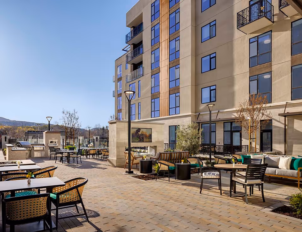 Outdoor patio area at Belmont Village Senior Living San Ramon featuring multiple seating arrangements with tables and chairs, a built-in fireplace with a mounted TV above it, modern lamp posts, and a multi-story building with large windows in the background under a clear blue sky.