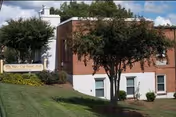 Exterior view of a brick building with white trim, surrounded by green grass and trees under a partly cloudy sky. A sign in front reads 'White Oak Manor York'.