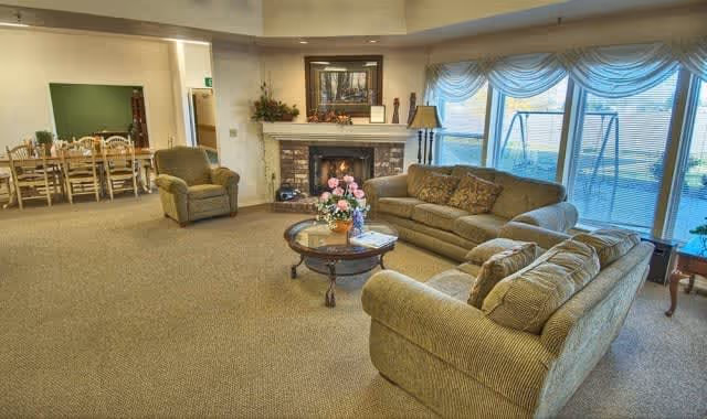 A cozy living room area with two sofas, an armchair, and a round coffee table with a flower arrangement on it. There is a fireplace with a painting above it and decorative items on the mantel. Large windows with sheer curtains let in natural light and show a swing set outside. In the background, there is a dining area with a wooden table and chairs.