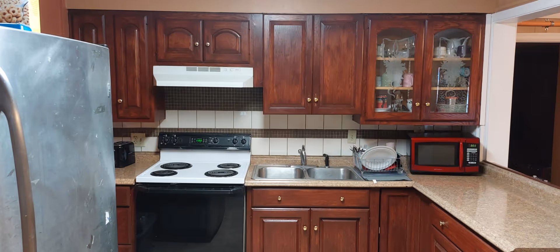 A kitchen with dark wooden cabinets, a white electric stove with a black oven, a double stainless steel sink, a red microwave on the countertop, and a stainless steel refrigerator on the left side. There are dishes drying in a rack next to the sink and various cups and containers visible through glass cabinet doors.