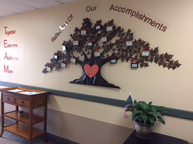 Interior wall display in a facility showing a large decorative metal tree with small plaques attached to its branches, titled 'Reflection of Our Accomplishments'. To the left, the word TEAM is vertically spelled out with the phrase 'Together Everyone Achieves More'. Below the display is a wooden table with papers on it and a potted plant with a small American flag on the floor nearby.