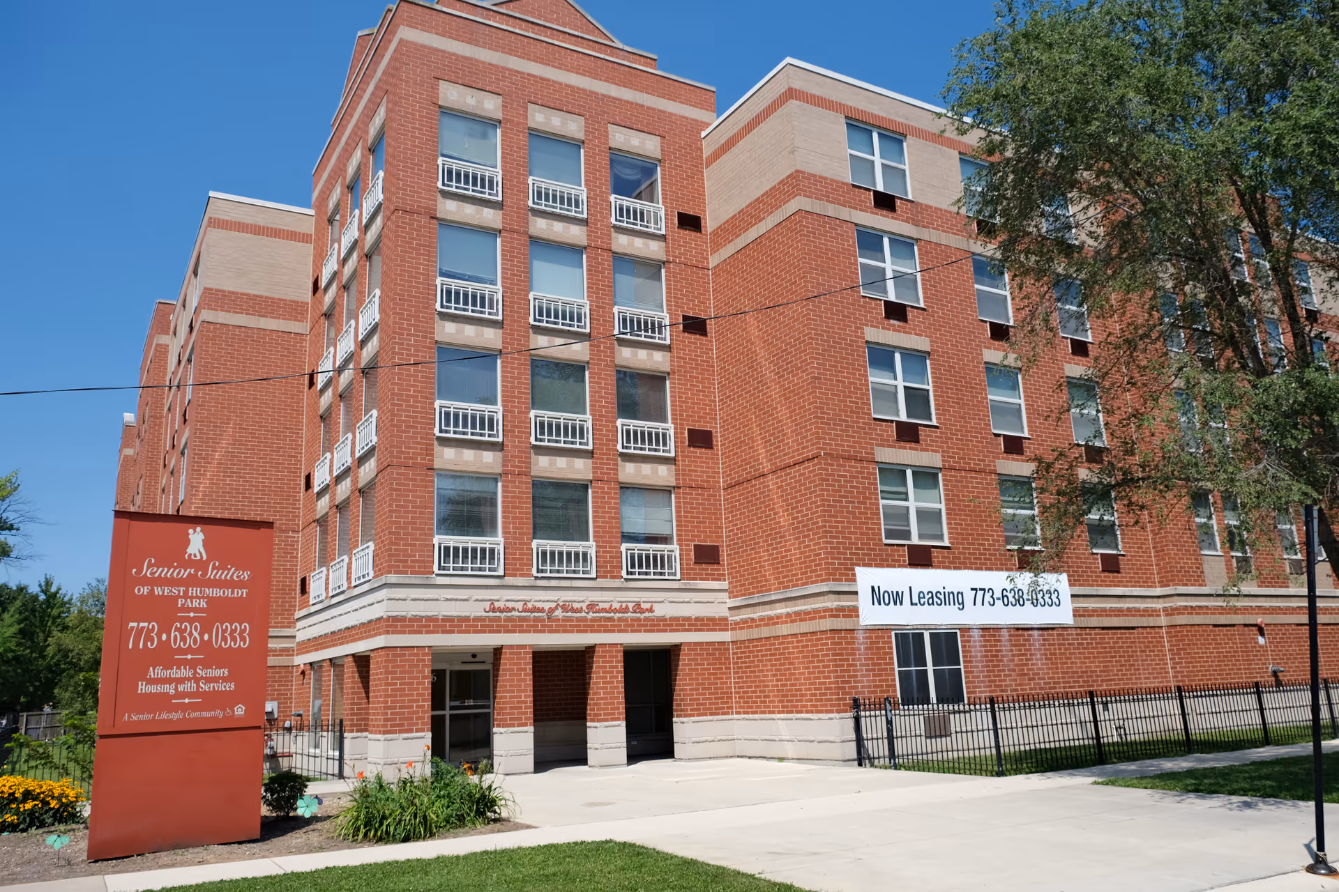 Exterior view of a multi-story brick building with multiple windows and a sign in front that reads Senior Suites of West Humboldt Park, Affordable Seniors Housing with Services. There is also a banner on the building that says Now Leasing with a phone number. The sky is clear and blue, and there are trees and grass around the building.
