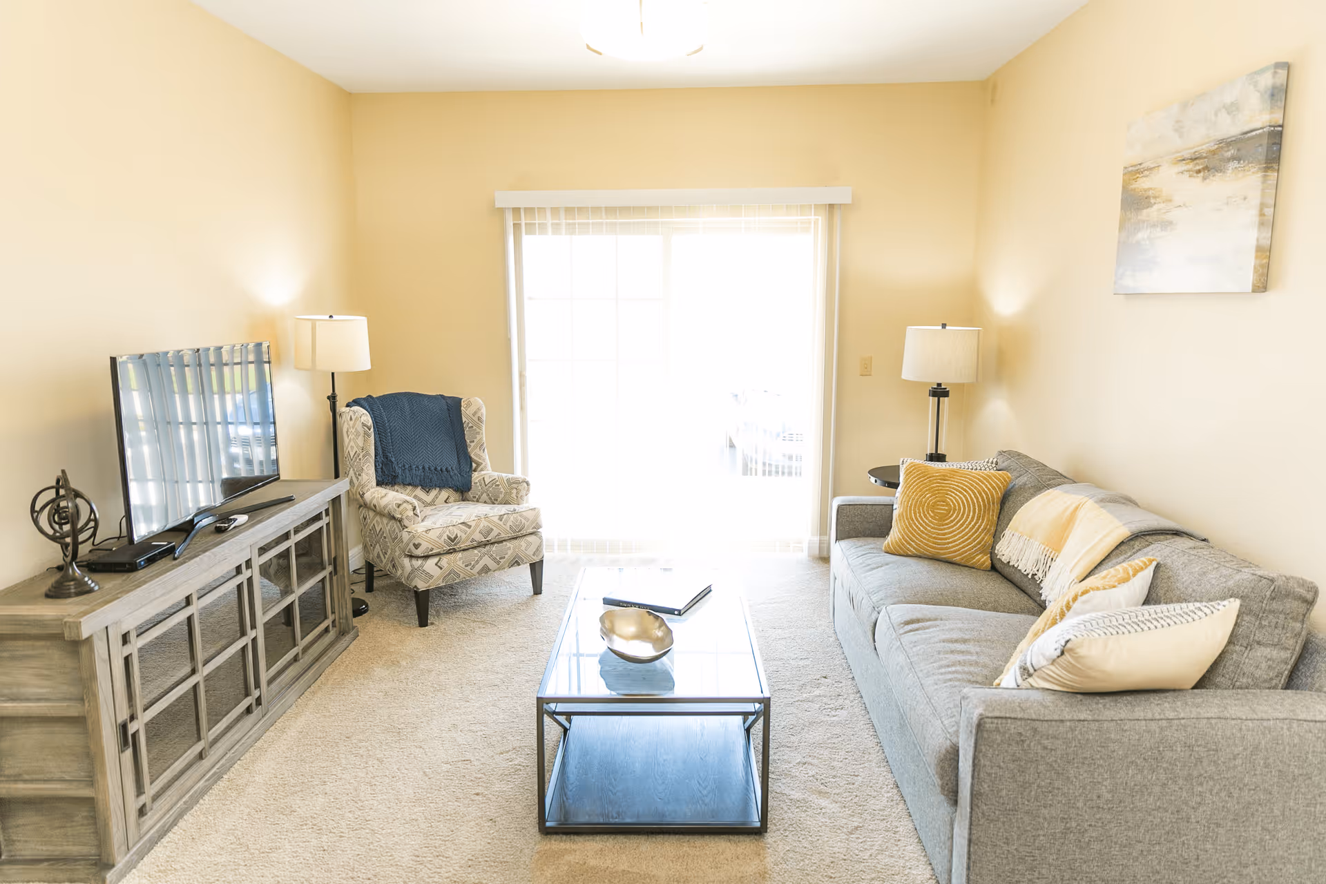 A bright living room with a gray sofa adorned with yellow and white pillows, a patterned armchair with a blue throw, a glass coffee table, a wooden TV stand with a flat-screen TV, two floor lamps, and a large window with vertical blinds letting in natural light.