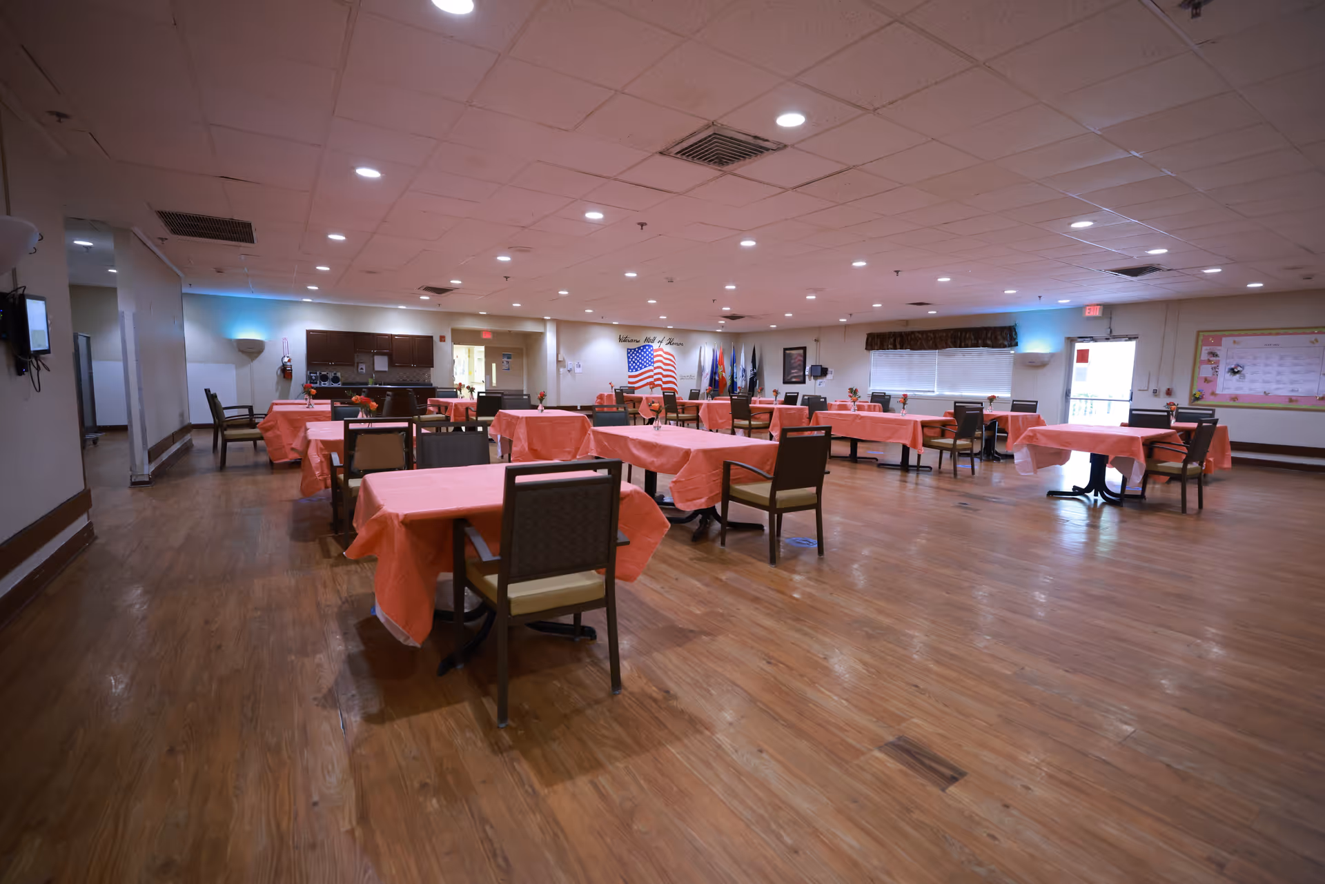 A spacious dining room with multiple tables covered in pink tablecloths and chairs arranged around them. The room has wooden flooring, a drop ceiling with recessed lighting, and an American flag displayed on the far wall. There are windows with blinds and a bulletin board on the right wall.