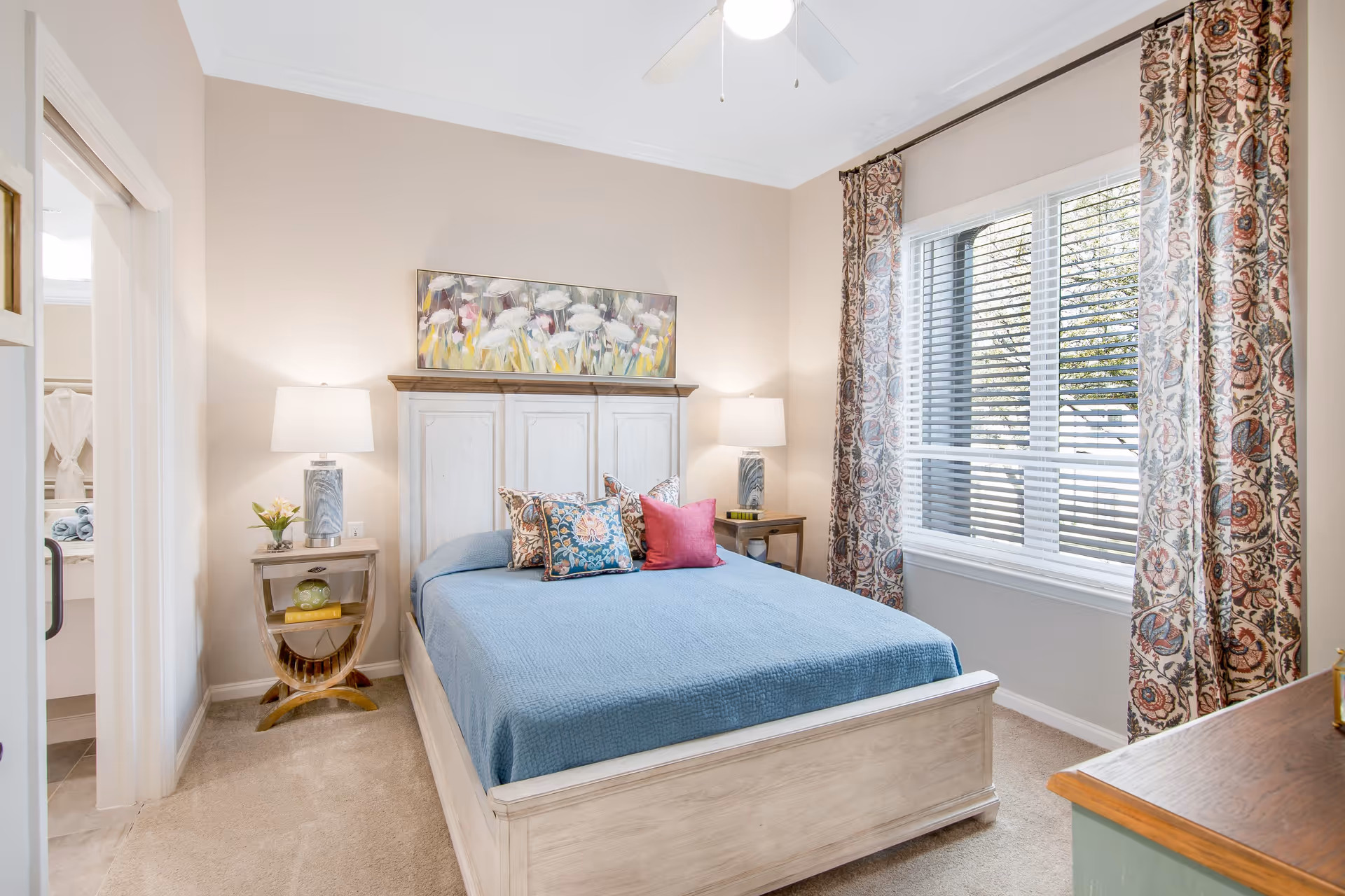 Sunlit, tastefully decorated bedroom with a bed in a blue coverlet, matching pillows, two bedside tables with lamps, a painting above the headboard, and a window with patterned curtains.