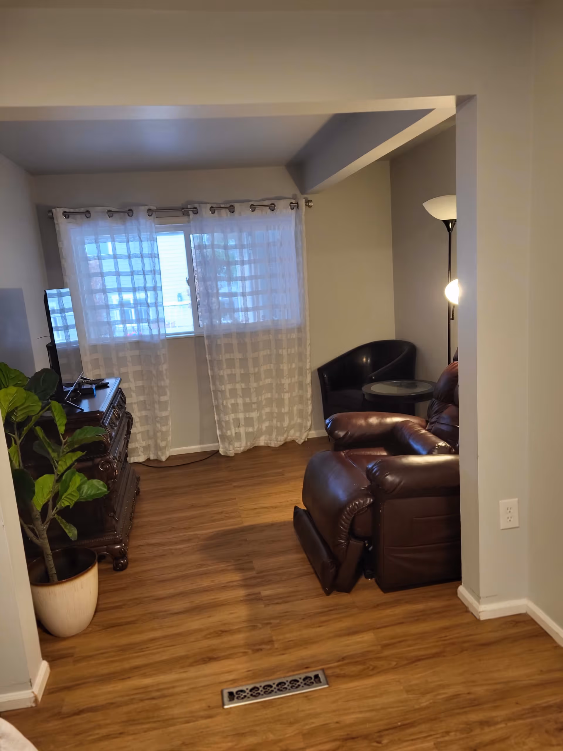 A cozy living room with wooden flooring, a large window with sheer white curtains, a dark brown leather recliner, a black armchair with a small round glass table beside it, a tall floor lamp, a wooden TV stand with a flat-screen TV, and a potted plant in the corner.