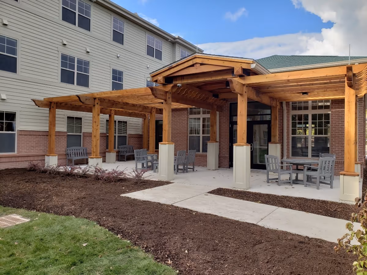 Outdoor patio area at Valley Bridge Apartments featuring wooden pergolas with seating arrangements including benches and chairs around tables. The building exterior has beige siding with brick accents and multiple windows under a partly cloudy sky.