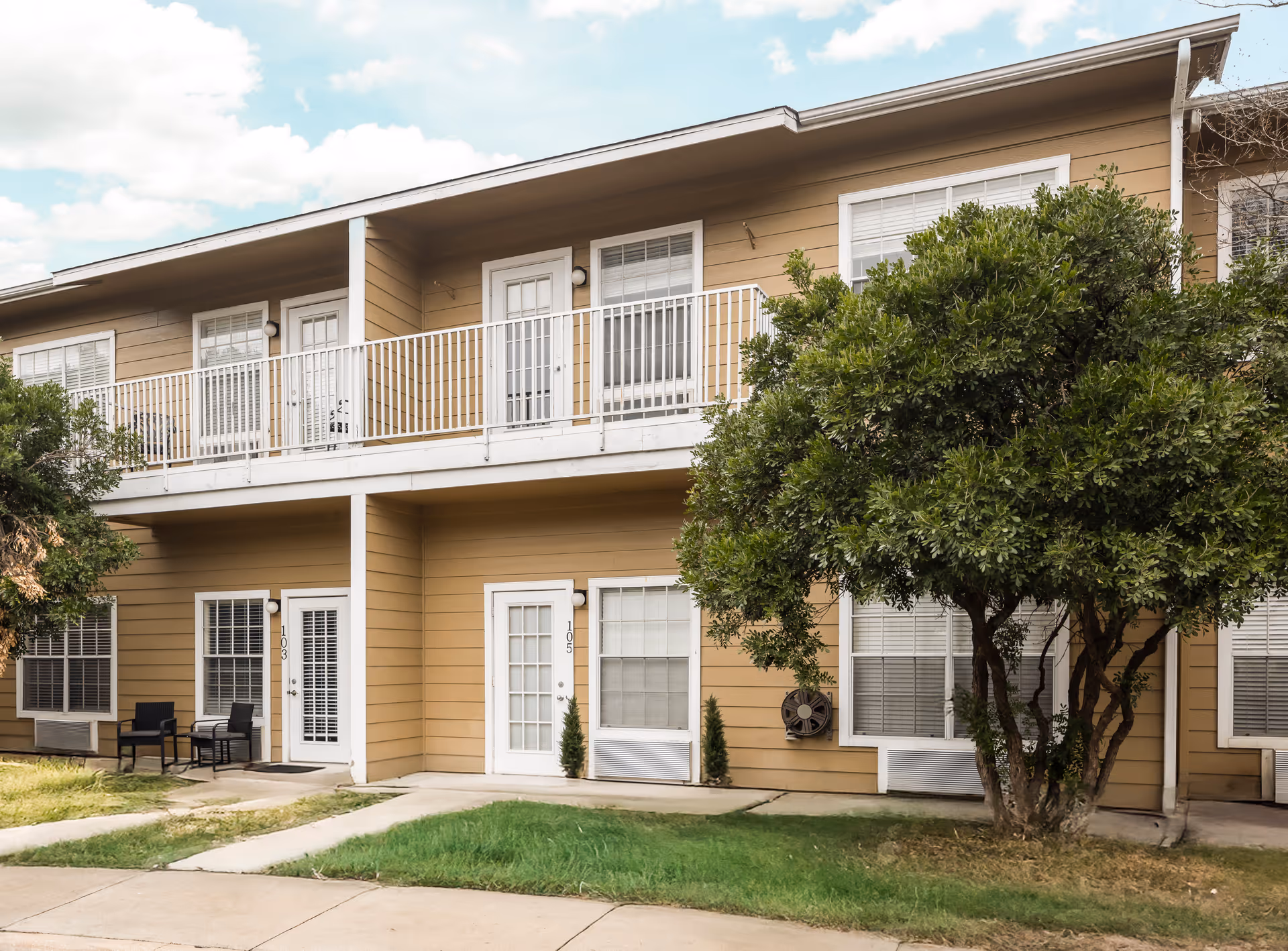 Exterior view of a two-story residential building with beige siding and white trim. The building has multiple doors and windows, a white railing on the upper balcony, and a small patio area with two chairs and a table. There is a green tree and grass in front of the building under a partly cloudy sky.
