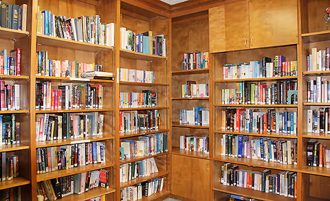 Interior view of a library or reading room with wooden bookshelves filled with a variety of books arranged neatly. The shelves cover the walls and there are cabinets above some sections.