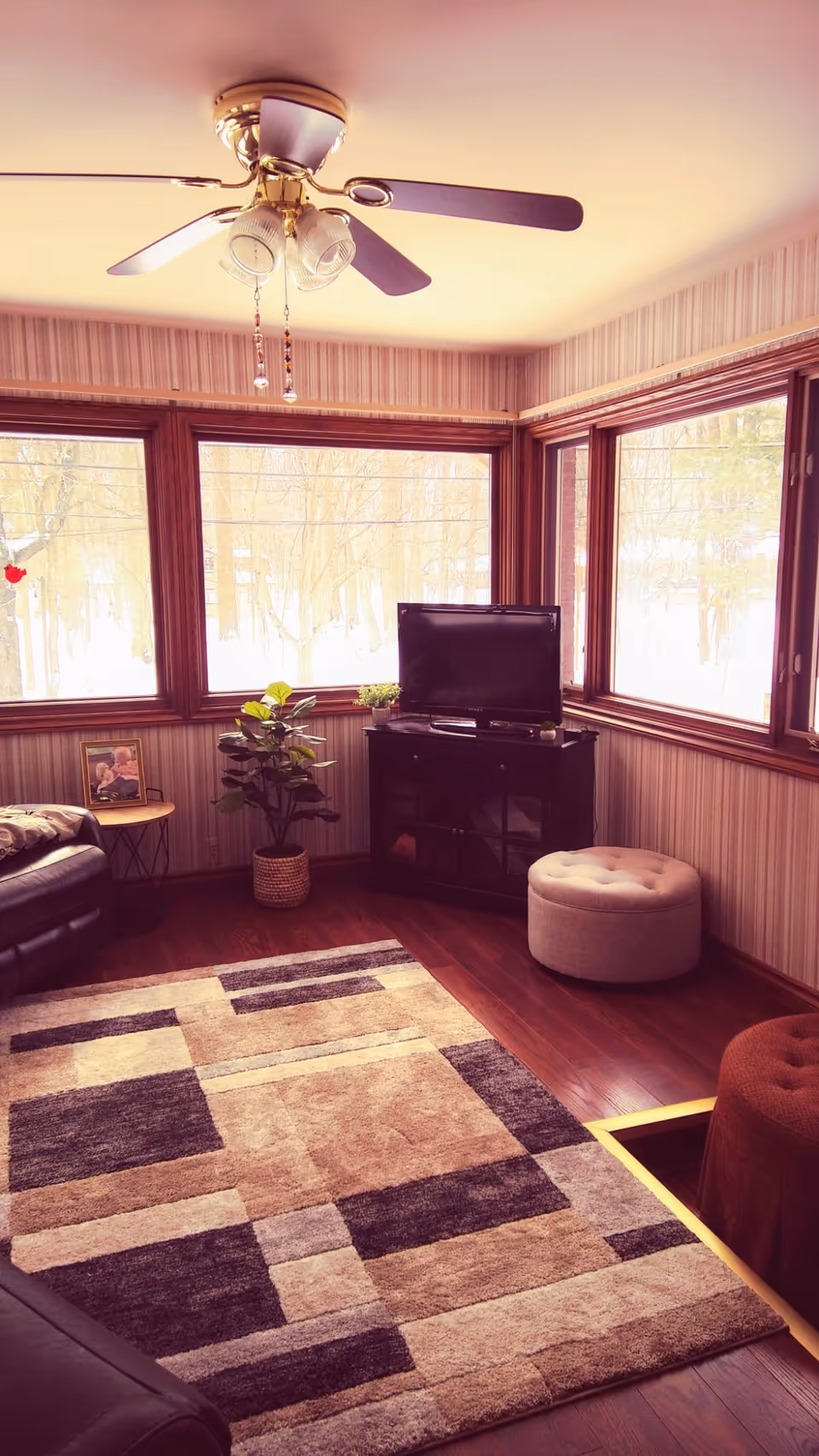 Cozy living room with large windows showing a snowy outdoor scene. The room features a ceiling fan with lights, a patterned area rug on wooden floor, a small TV on a black stand, a round beige ottoman, a potted plant, a side table with a framed photo, and leather seating partially visible.