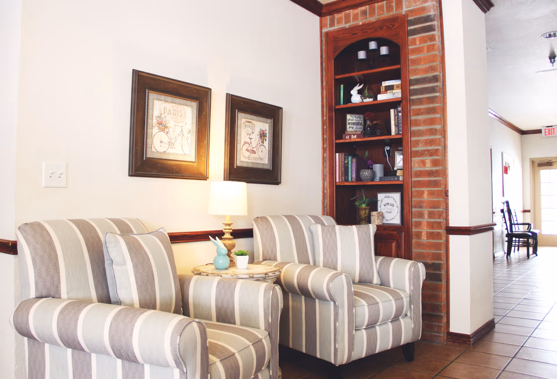 A cozy seating area with two striped armchairs and a small round table with a lamp and decorative items between them. Behind the chairs, there are two framed pictures on the wall and a built-in wooden bookshelf with books and decor. The area leads to a hallway with chairs and an exit door in the background.