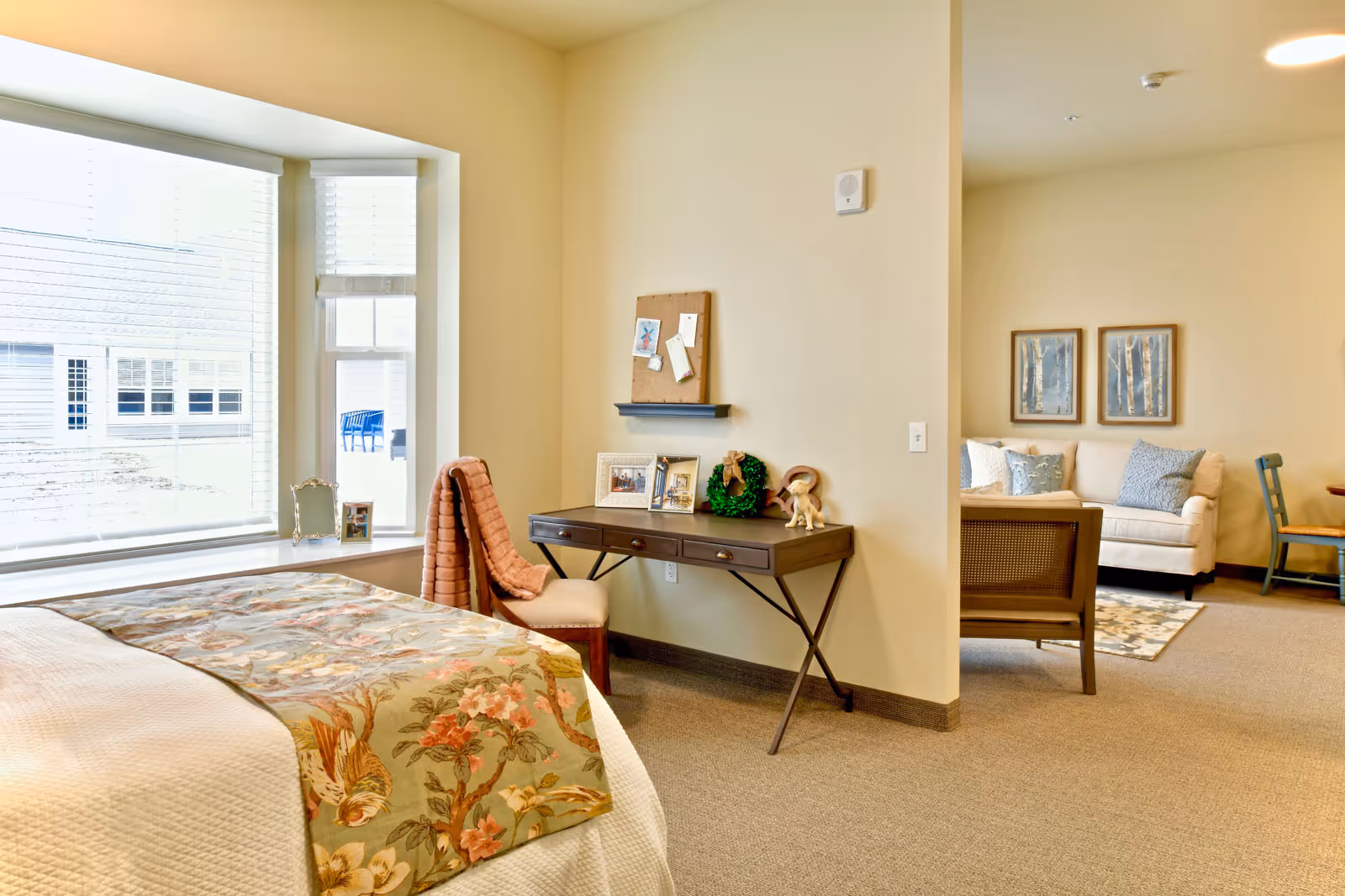A cozy senior living suite with a bed covered in a floral blanket near a large window with blinds. A wooden desk with three drawers holds framed photos, a small wreath, and a figurine, accompanied by a chair draped with a pink blanket. In the background, a sitting area features a beige sofa with blue and white pillows, two framed pictures on the wall, a wooden chair, and a small table.