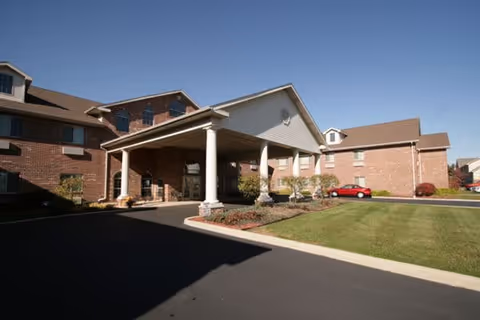 Exterior view of a senior living facility building with a covered entrance supported by white columns, brick walls, and a neatly maintained lawn and driveway.