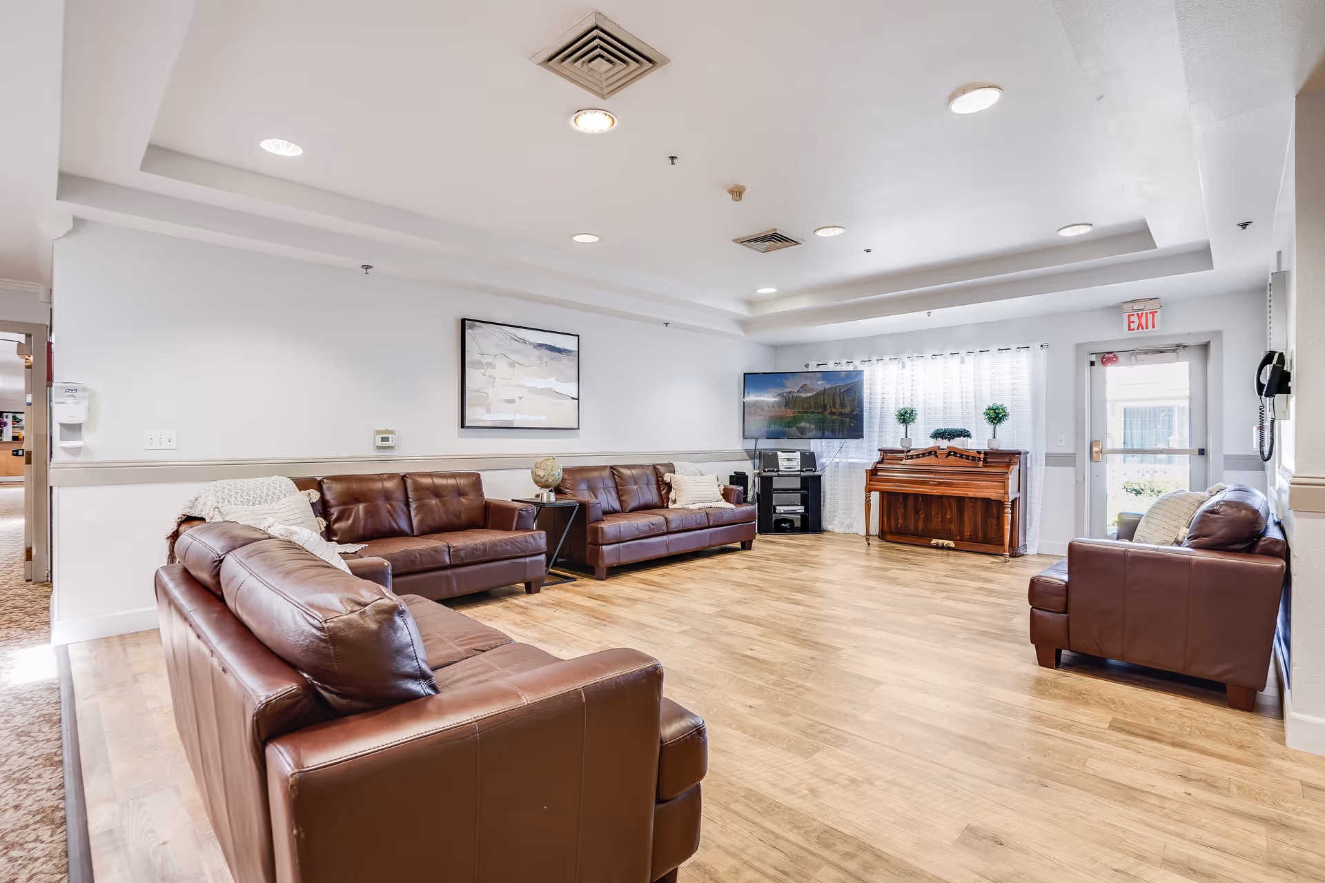 Bright communal living room with several brown leather sofas, a television, and an upright piano on wood flooring near an exit door.