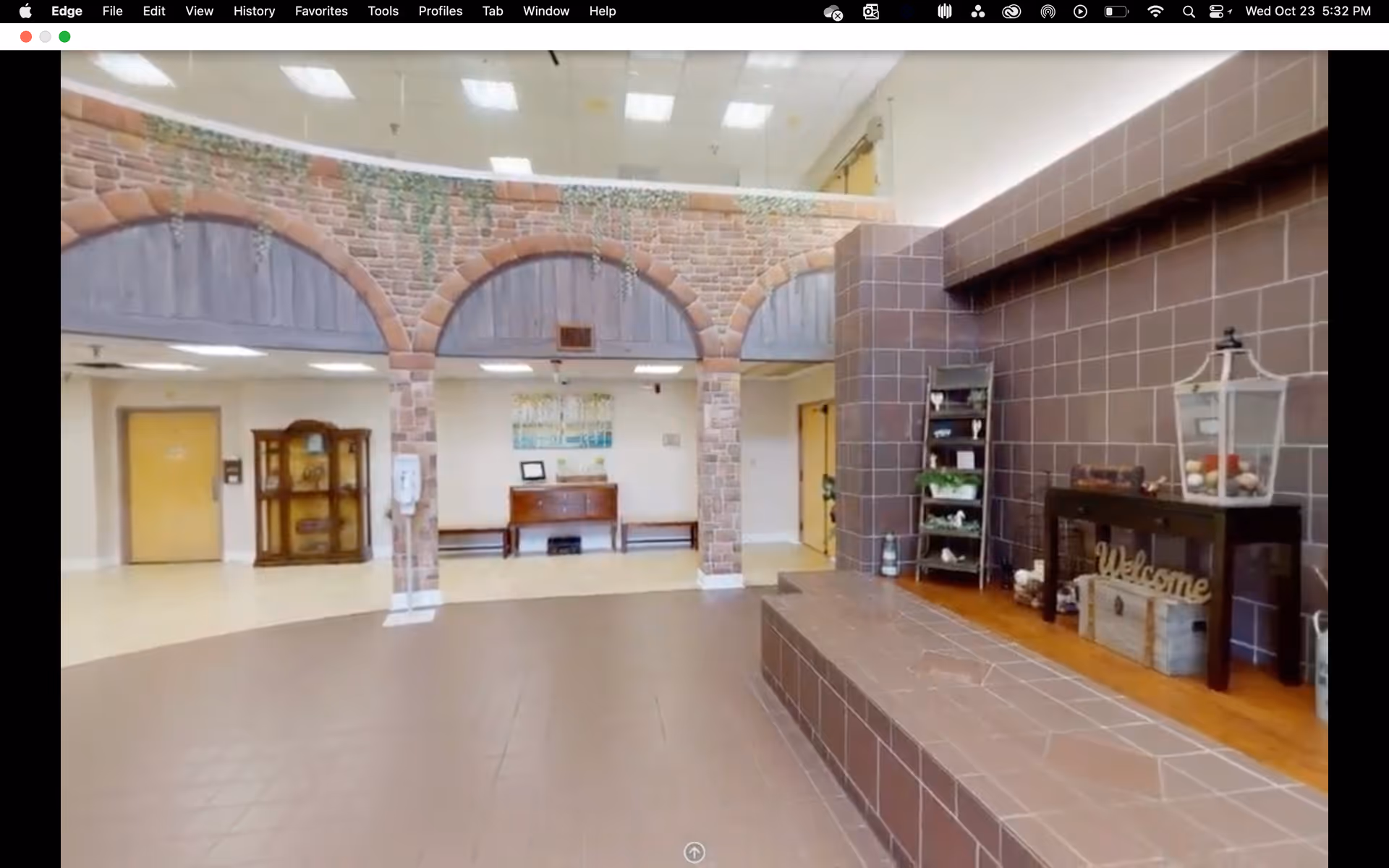 Interior view of a senior living facility lobby area with a decorative brick archway, a tiled raised platform with decorative items including a lantern and a 'Welcome' sign, a wooden cabinet with shelves holding plants and decor, and a bench against the far wall under a framed picture.