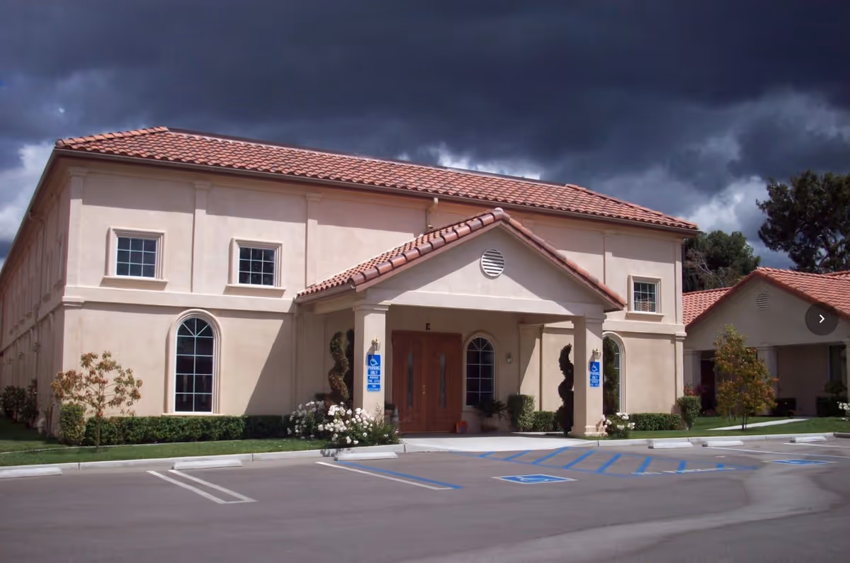 Front entrance of a two-story Mediterranean-style retirement building with a tiled roof, arched windows, landscaping, and marked handicap parking spaces.