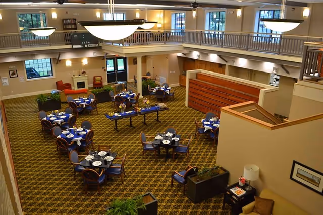 Interior view of a senior community dining room with multiple round tables set with plates, napkins, and utensils. The room has a patterned carpet, a fireplace with chairs nearby, large hanging light fixtures, and a second-floor balcony overlooking the area.