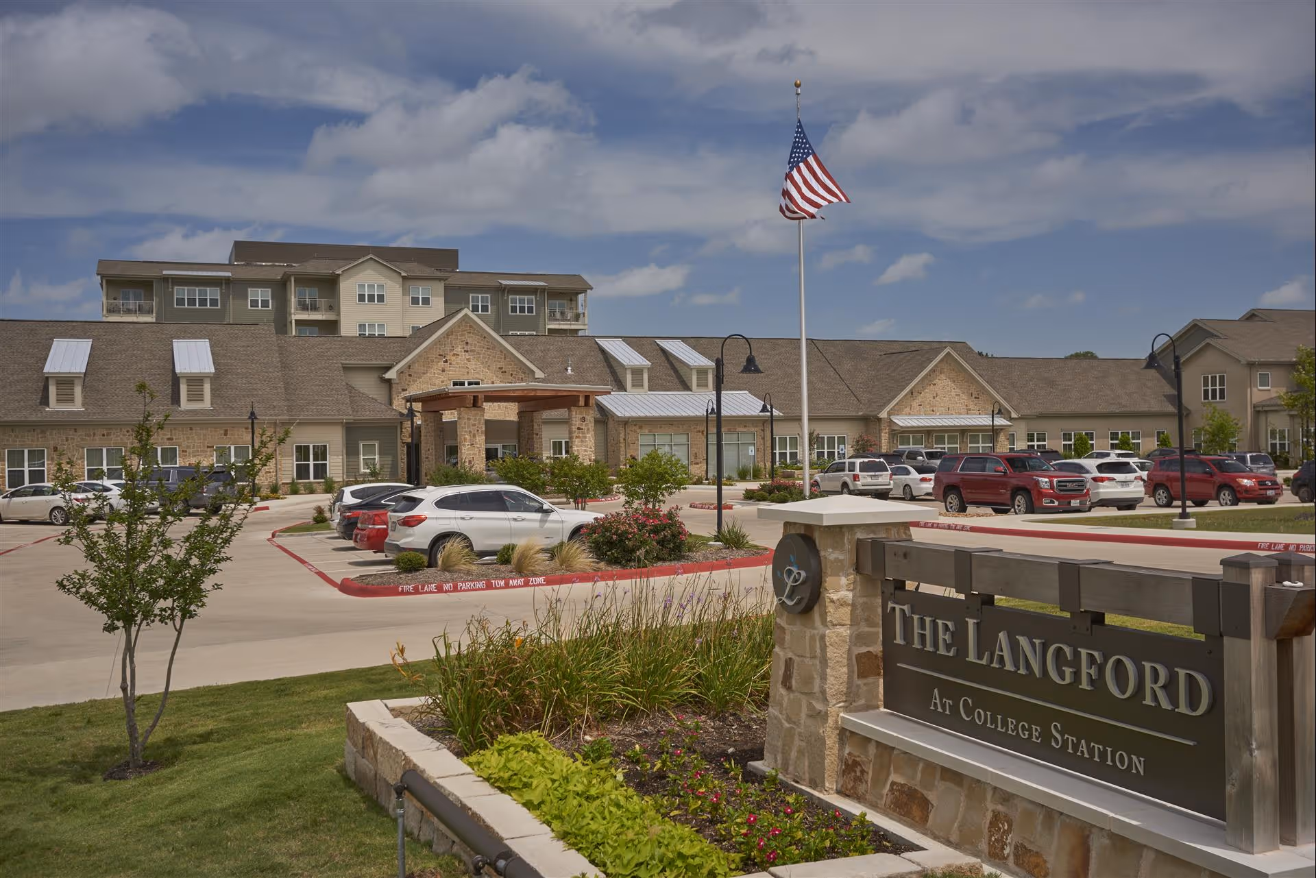 Exterior view of The Langford Methodist Retirement Community building with a parking lot in front, an American flag on a flagpole, landscaped greenery, and a sign that reads 'The Langford At College Station'.