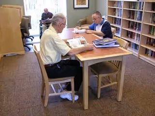 Two elderly men sitting at a wooden table in a room with bookshelves, reading and looking at documents. Another person is visible in the background sitting near a window. The room has carpeted flooring and framed pictures on the wall.