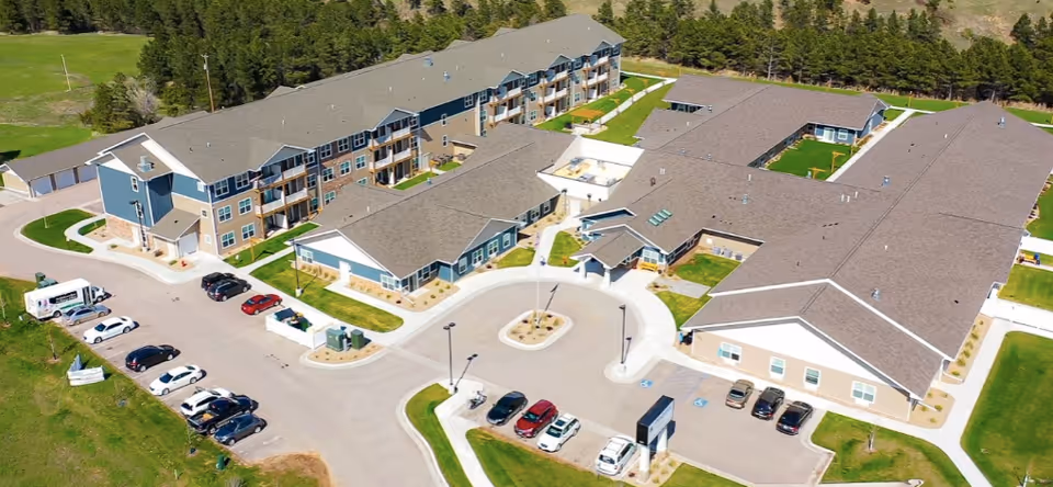 Aerial view of Peaceful Pines Senior Living at Rapid City showing multiple connected buildings with gray roofs, parking lots with cars, green lawns, and surrounding trees.