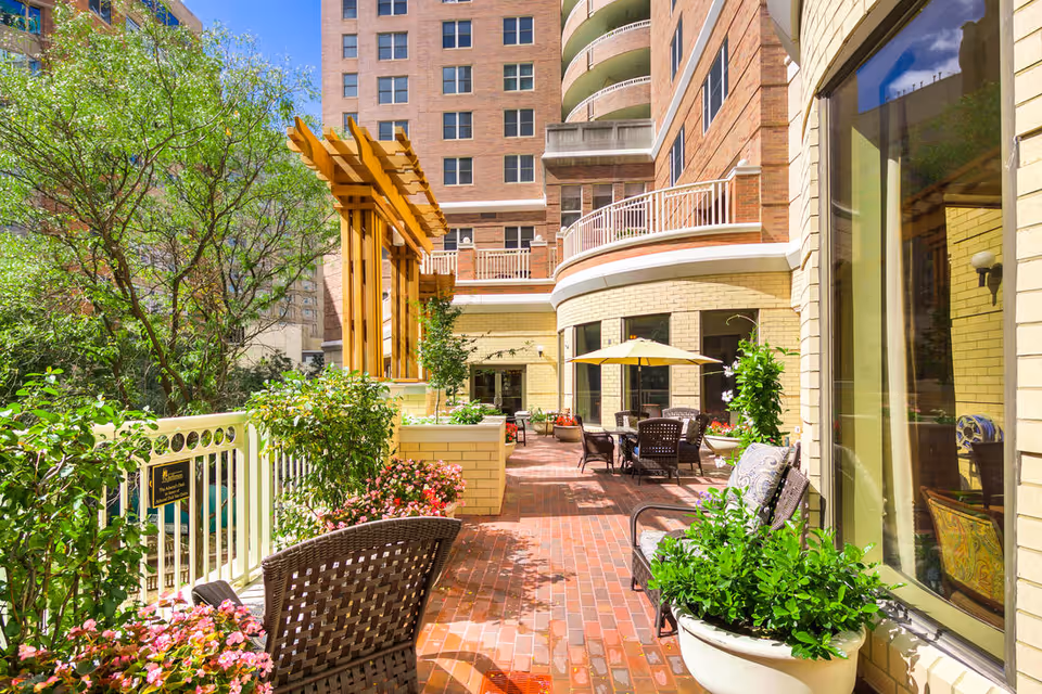 Outdoor patio area at The Jefferson with brick flooring, wicker chairs, tables with umbrellas, potted plants, and greenery. The patio is adjacent to a multi-story brick building with large windows and balconies.