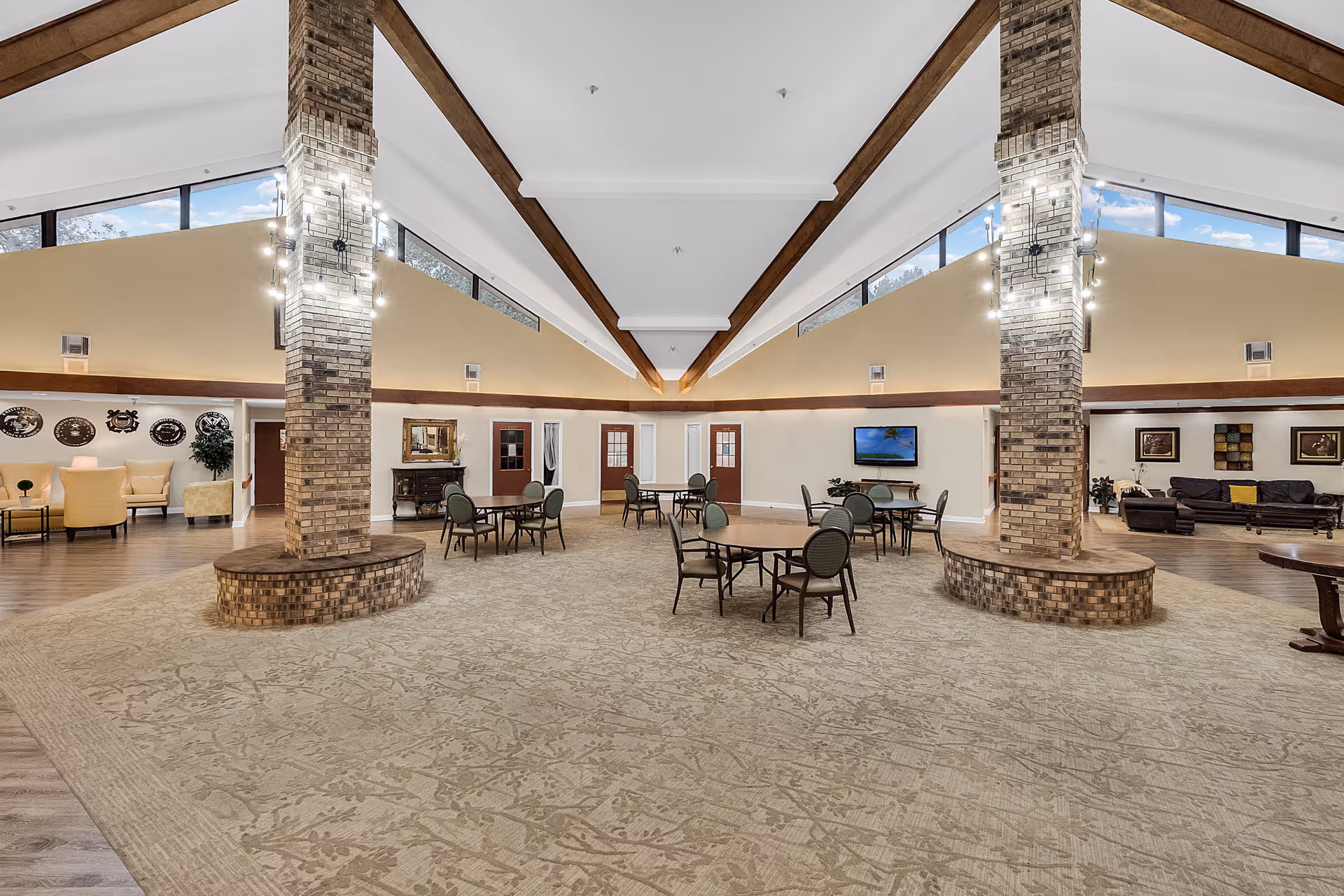 Spacious senior living facility common area with high vaulted ceilings, large windows near the ceiling, two brick columns with modern light fixtures, several round tables with chairs, a seating area with armchairs on the left, and a lounge area with sofas and framed artwork on the right.