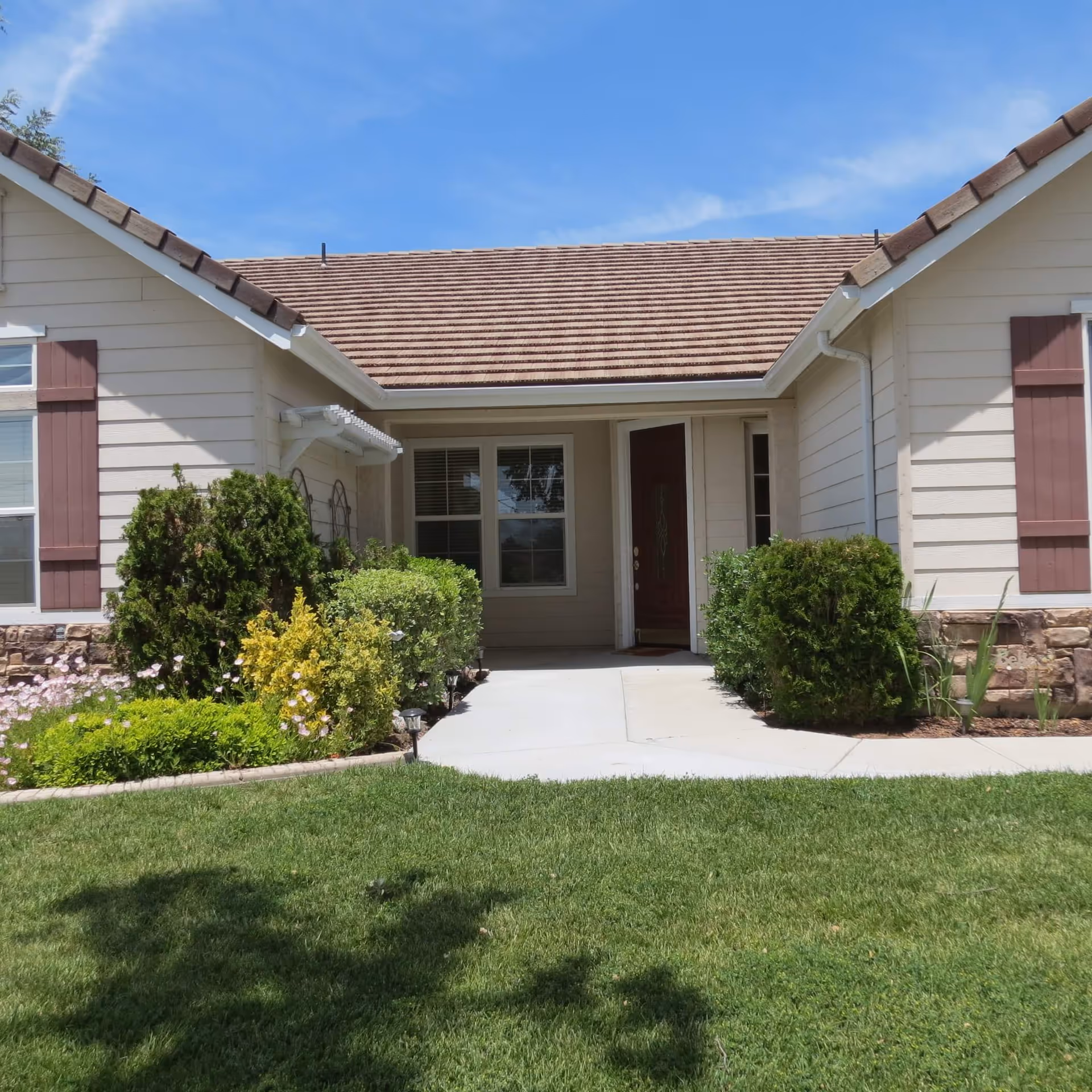 Front exterior view of a single-story house with beige siding, brown roof shingles, and a red front door. The entrance is framed by green bushes and a well-maintained lawn under a blue sky.