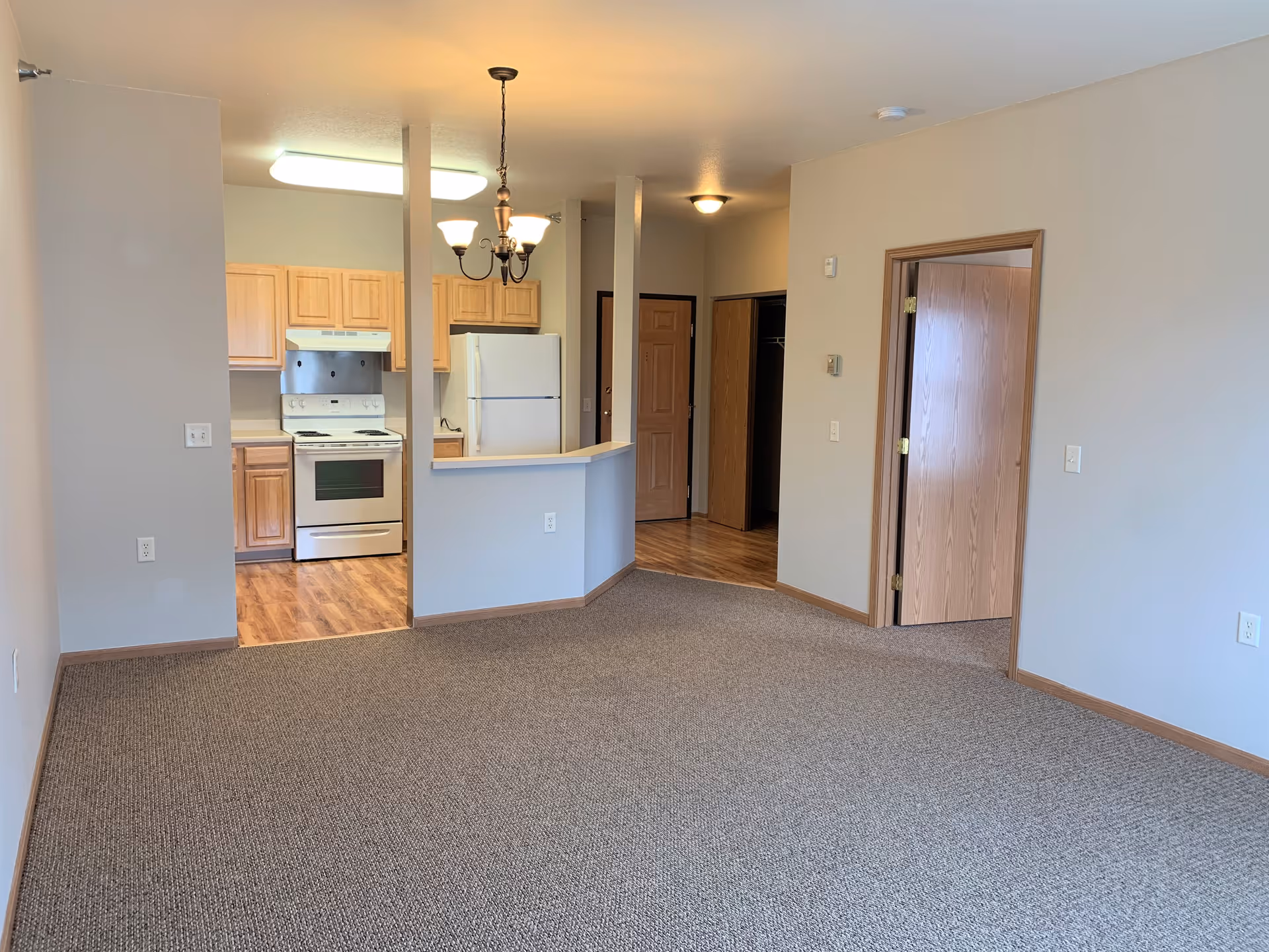 Carpeted living room open to a kitchen with light wood cabinets, white appliances, and a small breakfast bar.