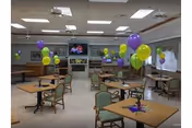 Communal dining/activity room with tables and chairs decorated with purple and yellow balloons and a TV above a fireplace.