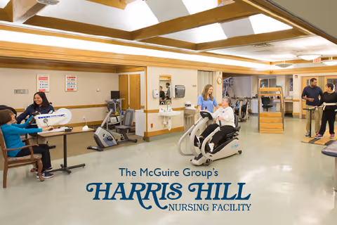 A spacious rehabilitation room in a nursing facility with several elderly individuals engaged in physical therapy activities using exercise equipment. A therapist is assisting a seated elderly woman on a stationary exercise bike, while others use different exercise machines and equipment. The room has wooden beams on the ceiling and light-colored walls.