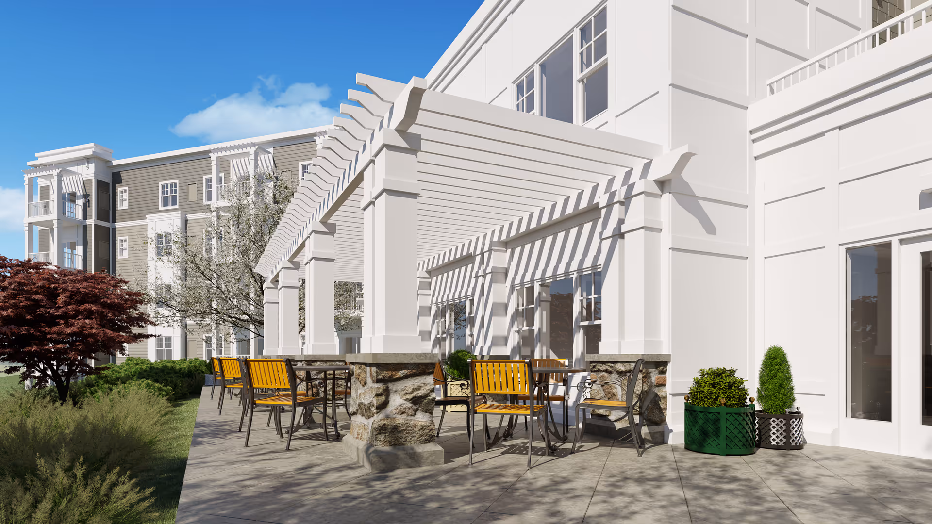 Outdoor patio area at a senior living facility with several tables and yellow chairs under a white pergola attached to a white building. There are plants and trees surrounding the patio, and a multi-story building is visible in the background under a clear blue sky.
