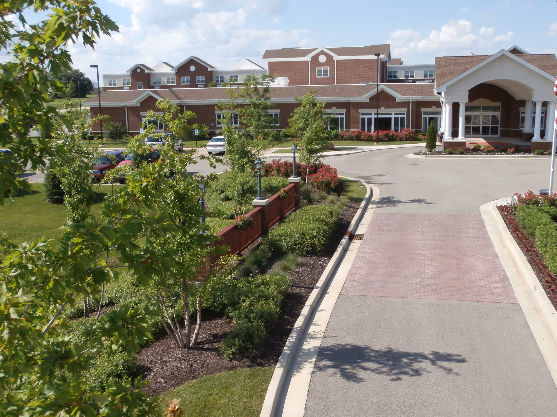 Front entrance and driveway of a brick senior living facility with a porte-cochère, landscaped gardens, and trees.