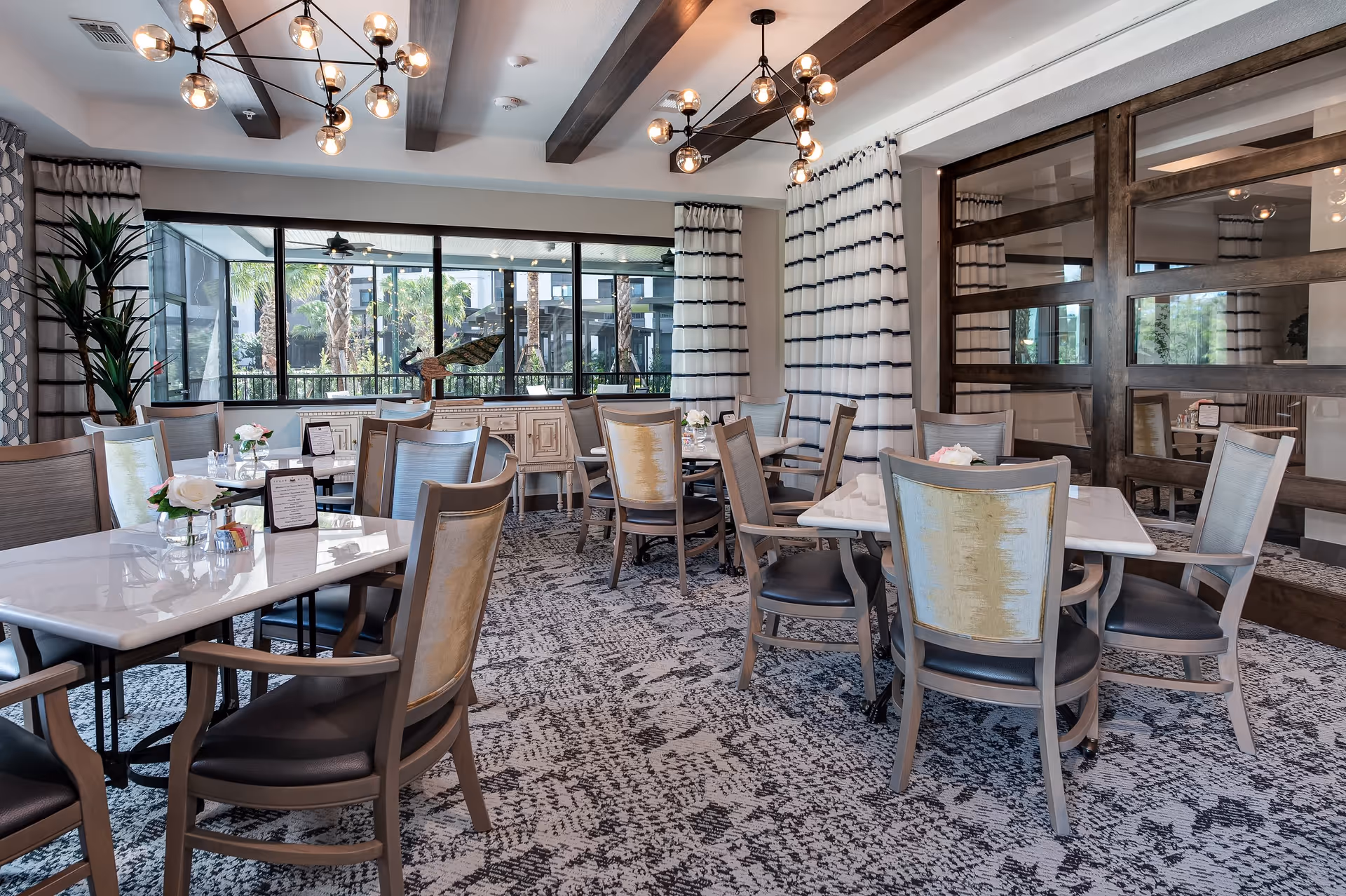 Bright, modern dining room with multiple tables and chairs, large windows overlooking greenery, patterned carpet, and pendant lighting.