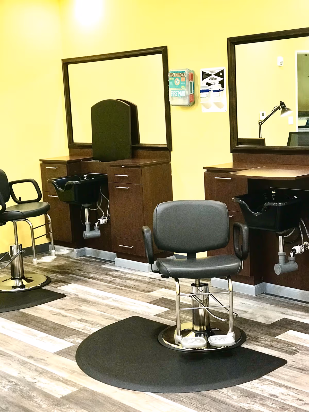 Interior of a salon area with two black salon chairs in front of wooden cabinets with mirrors. Each station has a black sink for washing hair. The walls are painted yellow and the floor has a wood-like pattern. A first aid kit is mounted on the wall between the mirrors.