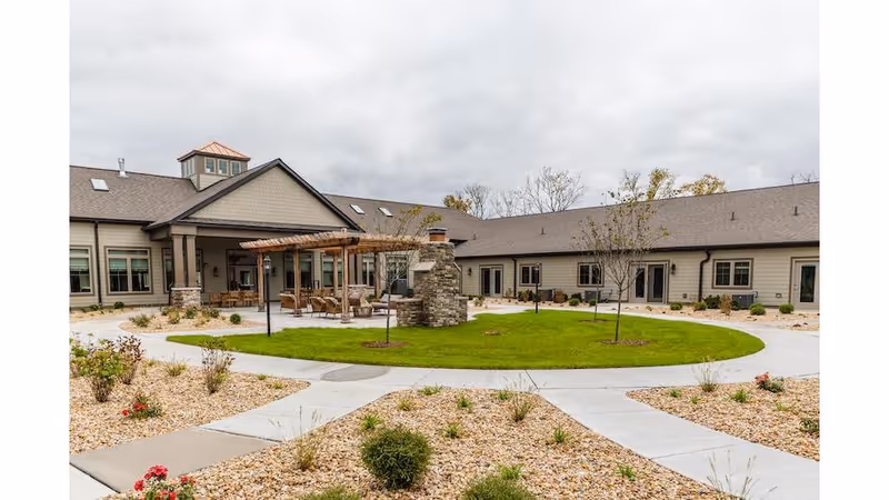 Outdoor courtyard area of Cedarhurst Senior Living of Frankfort featuring a circular green lawn with a stone fireplace in the center, surrounded by a paved walkway and landscaped with small bushes and rocks. The building surrounds the courtyard with multiple windows and a covered patio area with seating.