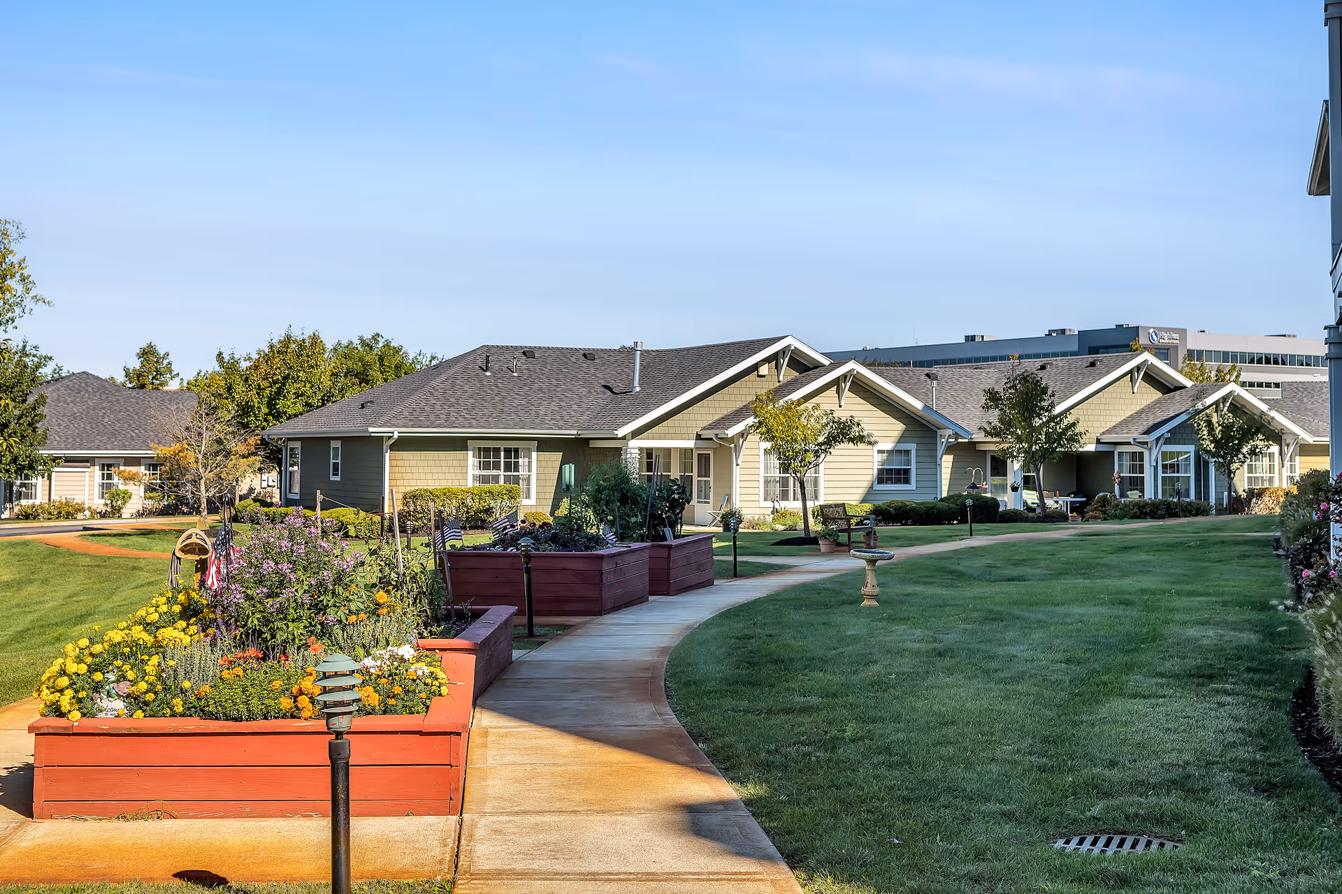 A curved walkway winds past raised flower beds and a lawn toward single-story residential buildings under a clear blue sky.