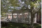 Outdoor view of a white wooden gazebo surrounded by trees and grass, with a picnic table nearby and a single-story building in the background under a clear sky.